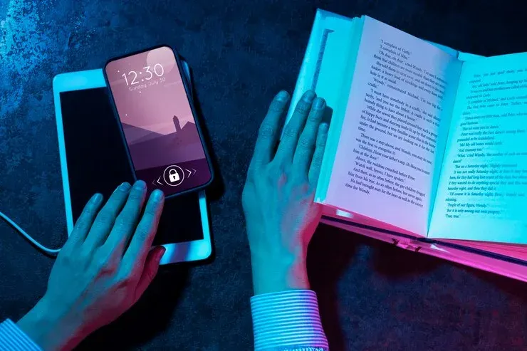 Hands on a smartphone, tablet and open book lit in neon blue