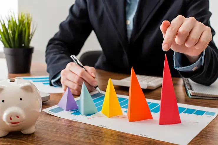 Person charting growth with colourful paper cones on a bar graph beside a piggy bank