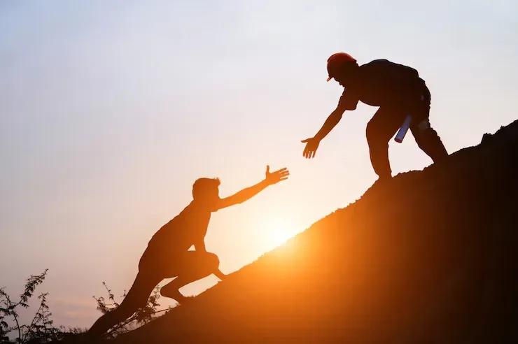 Silhouette of a person reaching down to help another climb a hill at sunset