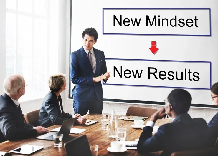 Presenter at a meeting beside a board reading New Mindset leads to New Results
